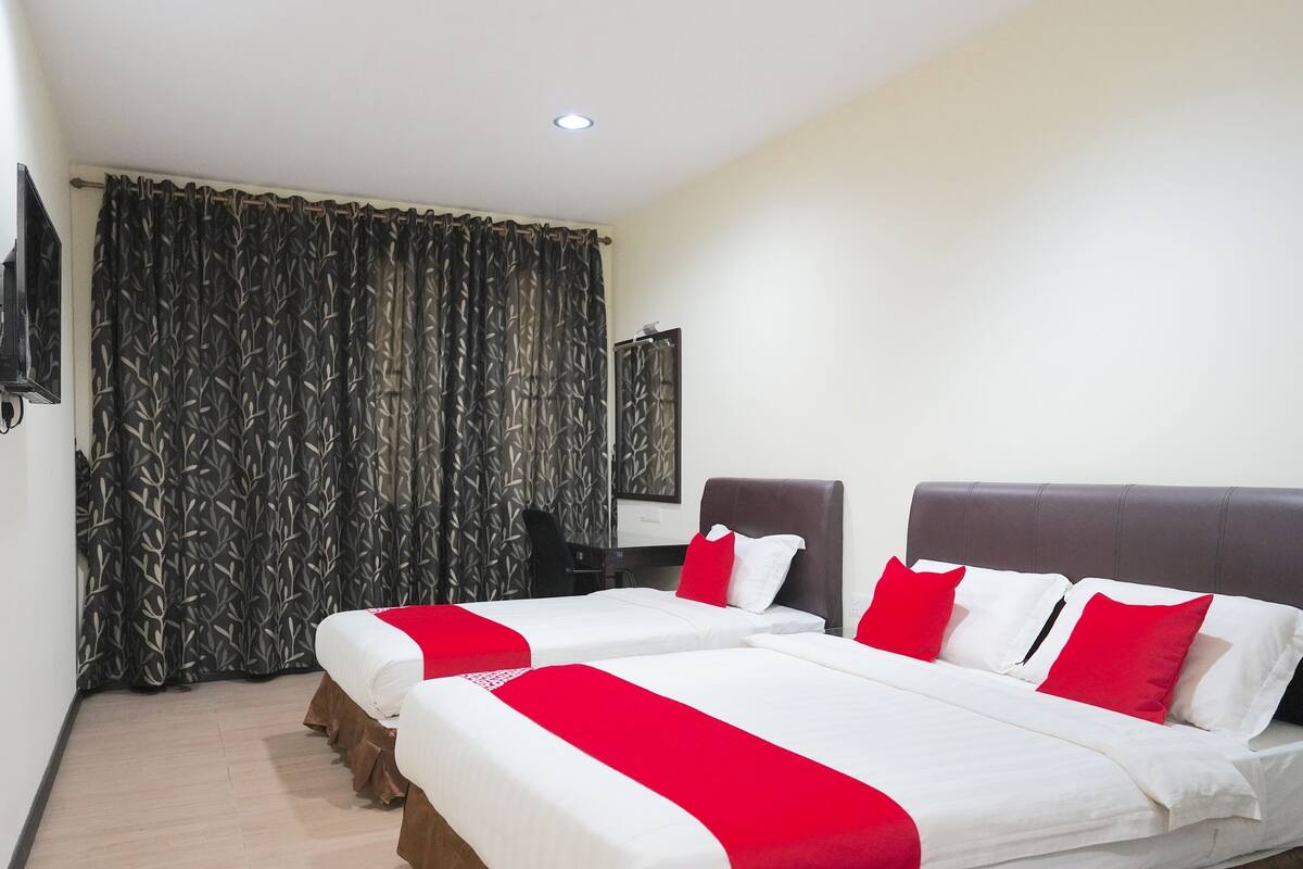 A hotel room with two beds, each with white bedding and red accents, a large window with patterned curtains, a desk, and a mirror.