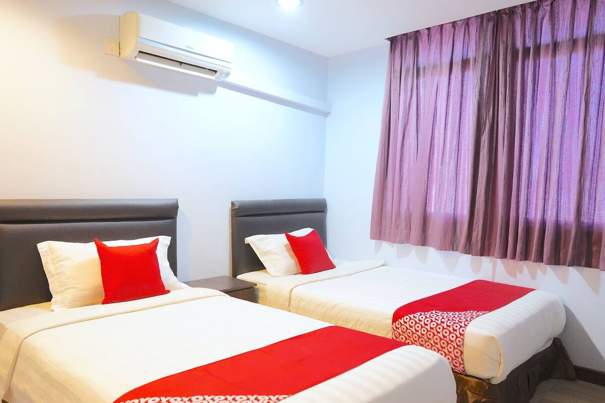 A hotel room with two beds, each with a headboard, a red and white bedspread, and a patterned blanket. There is an air conditioning unit on the wall and a window with purple curtains.