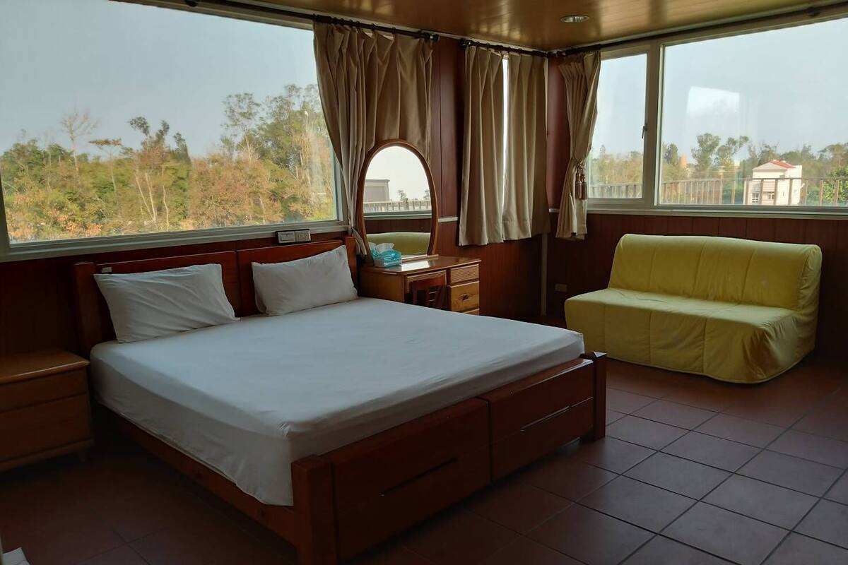 A bedroom with a large bed, a bedside table, a nightstand, a yellow sofa, and a window with curtains.