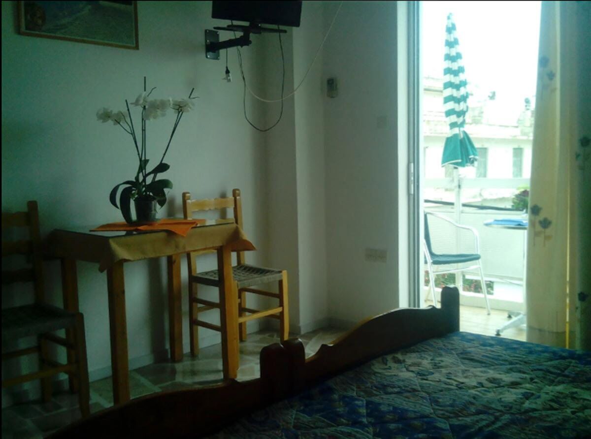 A bedroom with a bed, a wooden chair, a small table with a vase of flowers, and a view of the outside balcony.