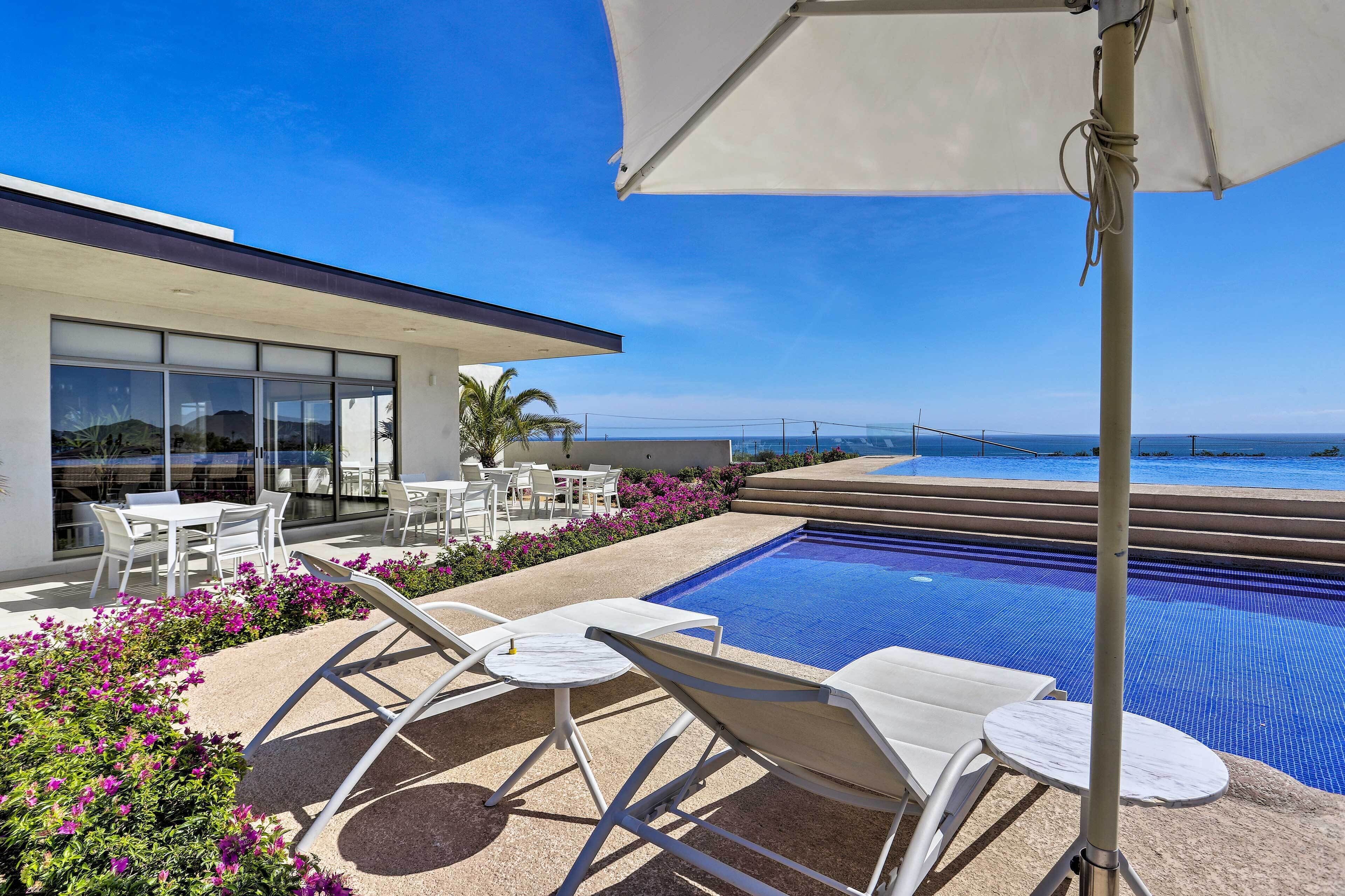 A modern outdoor pool area with lounge chairs and tables, a clear view of the ocean, and a building with large windows.