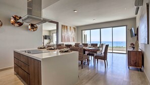 Una cocina moderna con una isla central, comedor con sillas de mimbre, ventanas amplias con vistas y un televisor de pantalla plana montado en la pared.