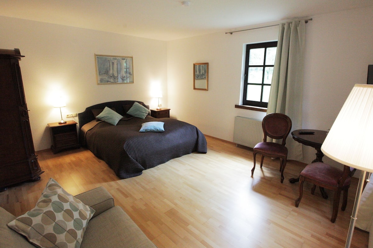 A bedroom with a bed, two bedside tables, a chair, a small table, and a window with curtains.