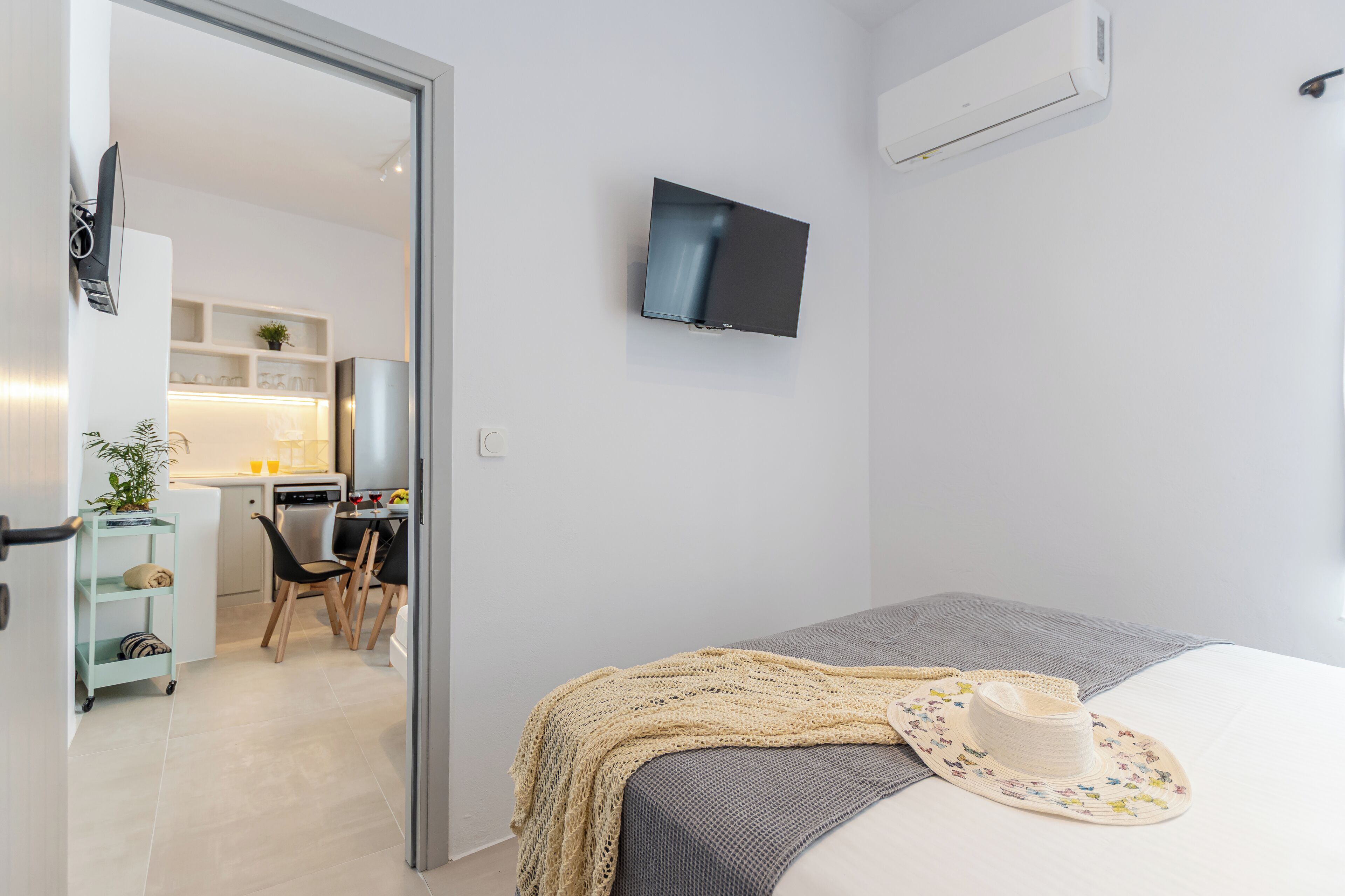 A modern hotel room with a bed, a TV mounted on the wall, a kitchenette, and a dining area.