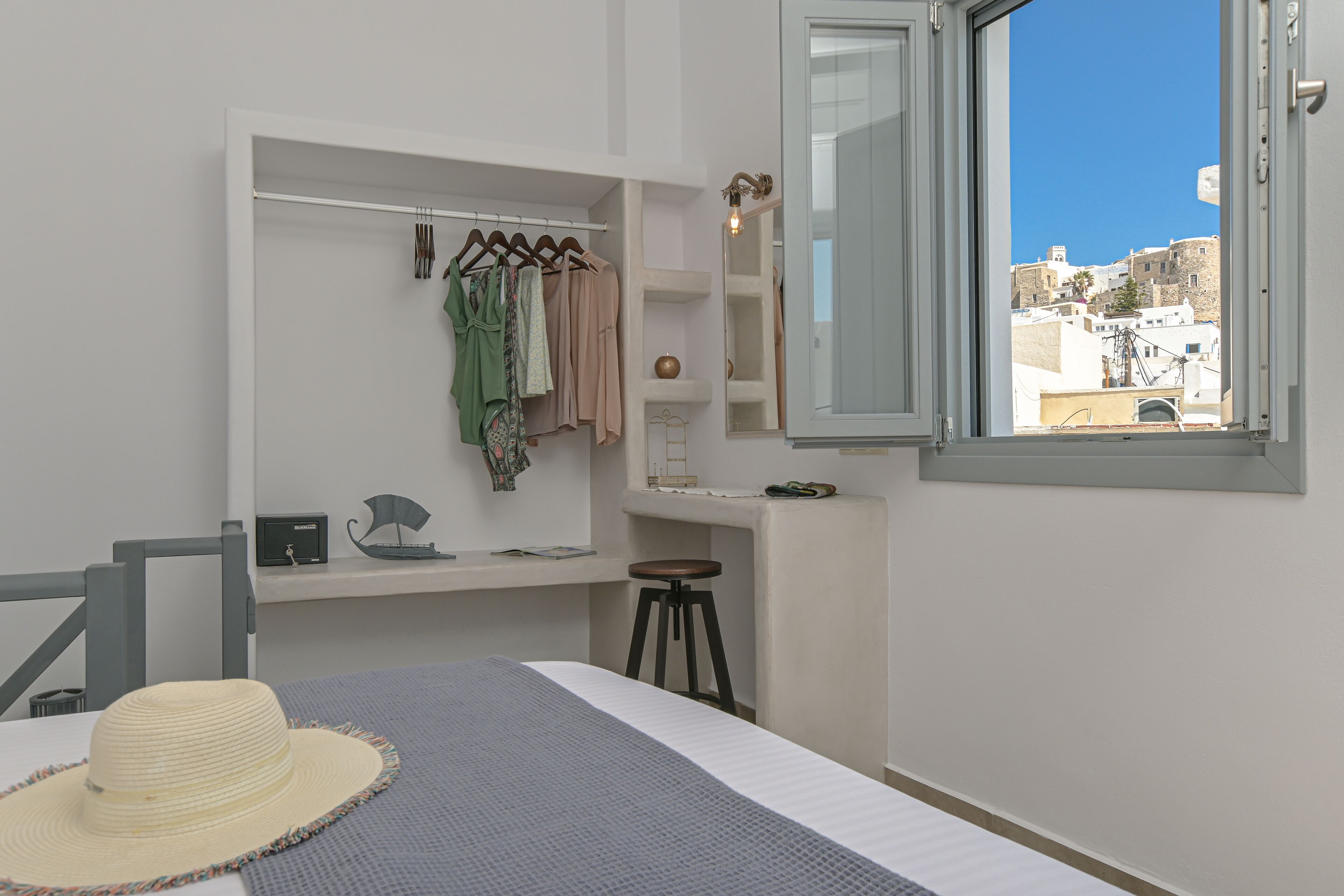 Een slaapkamer met een bed, een hangstoel, een krukje, een kledingkast en een raam met uitzicht op gebouwen.