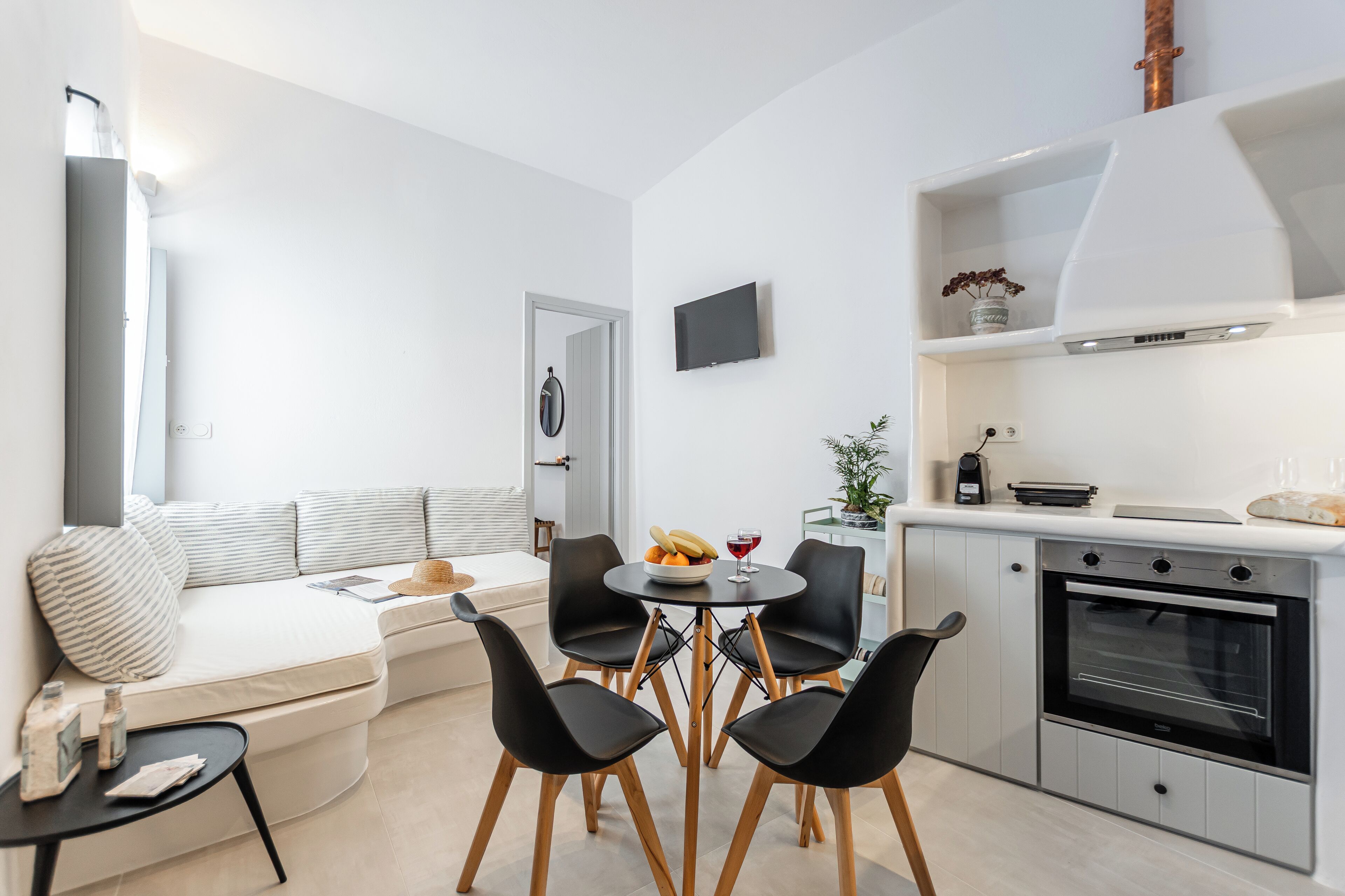 A modern kitchen with a built-in oven, a dining area with a round table and four chairs, and a seating area with a sofa and cushions.
