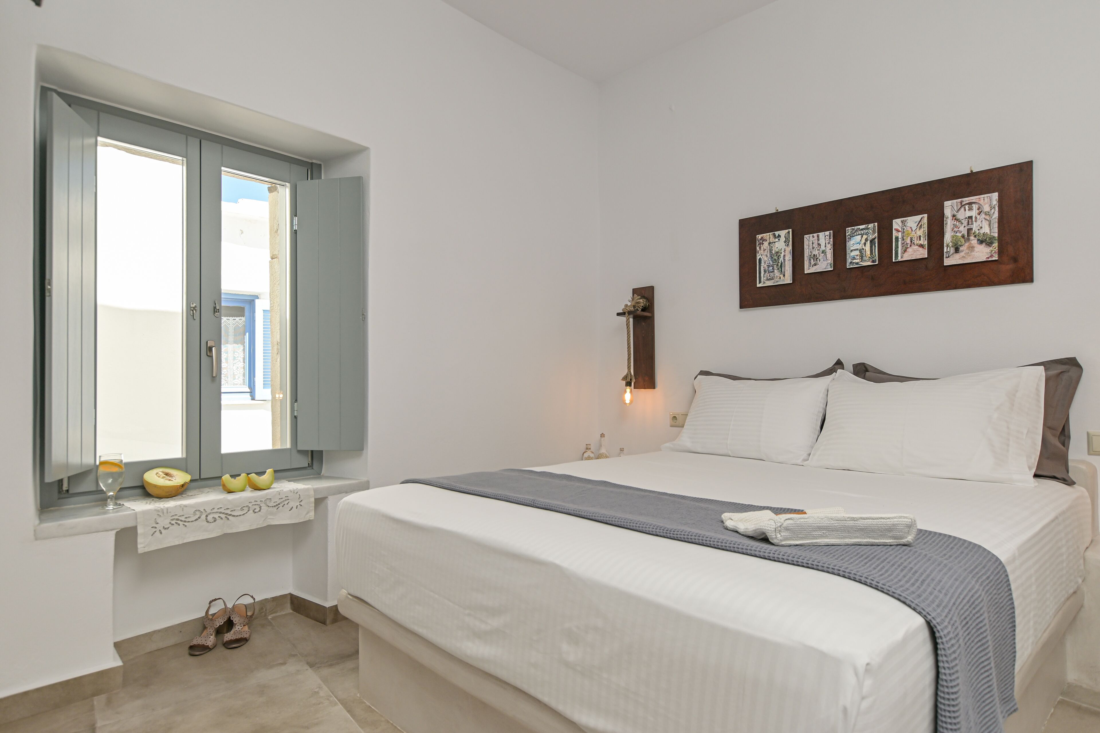 A bedroom with a bed, a window with open shutters, a wall-mounted artwork, and a bedside table with a lamp.