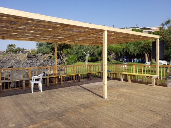 A wooden deck with a pergola, benches, and a view of a landscaped garden with trees and a rocky hillside.