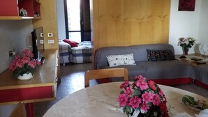 A cozy living room with a sofa, a dining table with a vase of flowers, and a bed with a checkered blanket.