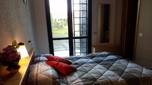 A bedroom with a bed, a bedside table with a lamp, and a window with a view of greenery.