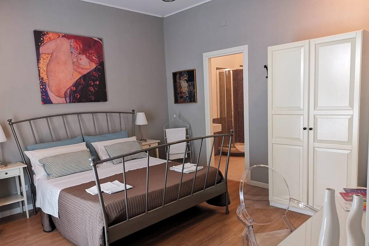 Un dormitorio con cama de estructura metálica, mesita de noche, lámpara y un cuadro en la pared. Hay una puerta que da a un baño con ducha.