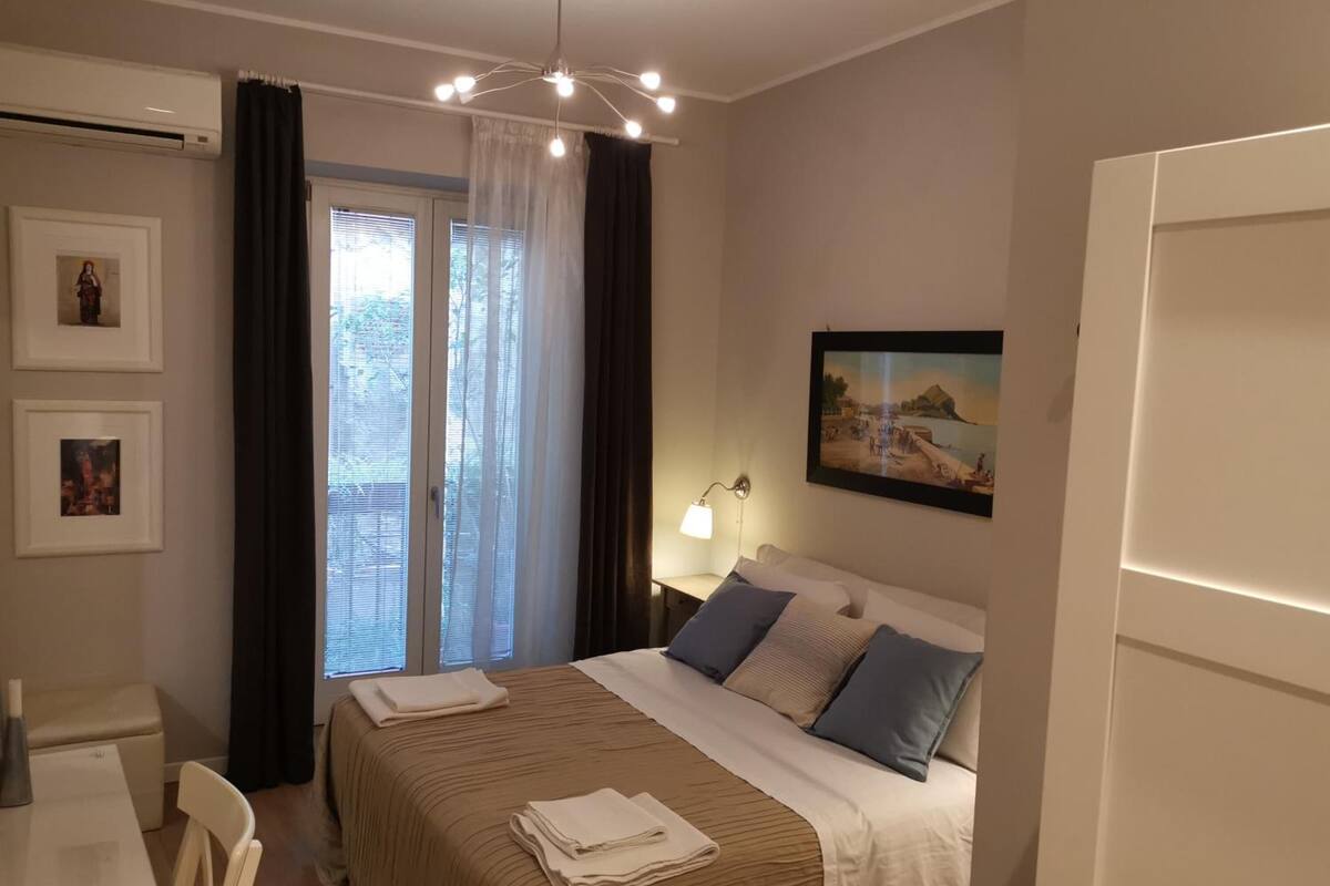 Una habitación de hotel con una cama, un escritorio, una silla, una ventana con cortinas, un cuadro en la pared y una lámpara en el techo.