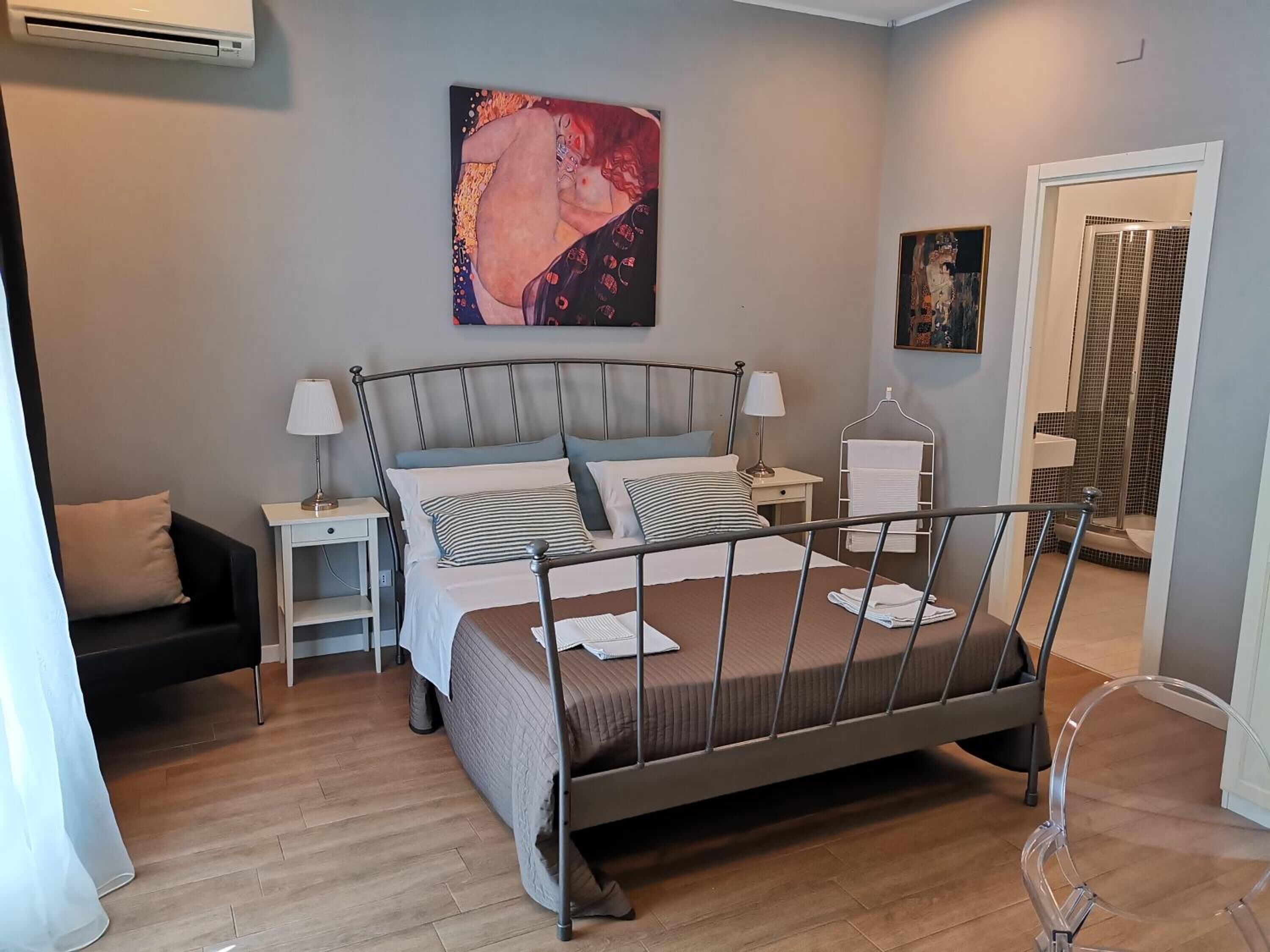 A modern bedroom with a metal bed frame, a bedside table with a lamp, a small shelf, and a wall-mounted painting.