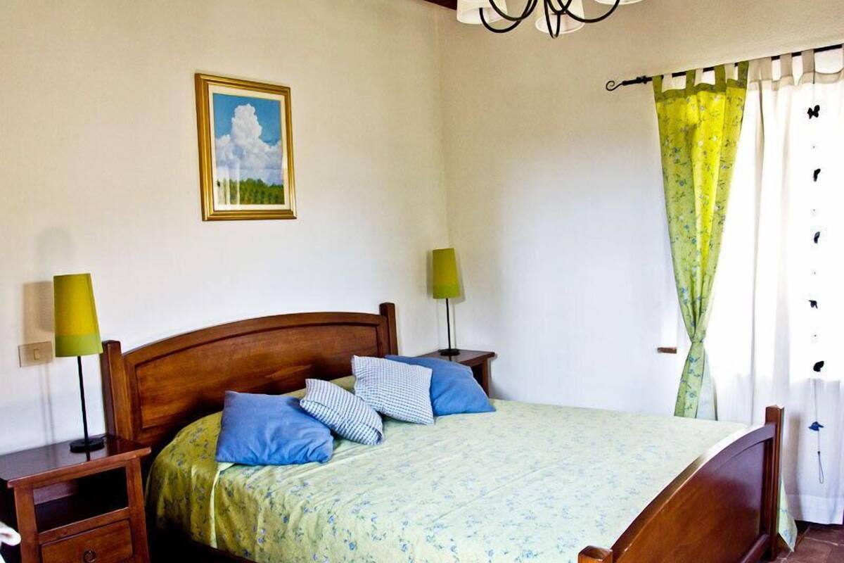 Une chambre avec un lit en bois, deux tables de chevet, une petite table, une couvre-lit à motifs verts, un oreiller bleu, un tableau encadré au mur et un rideau vert.
