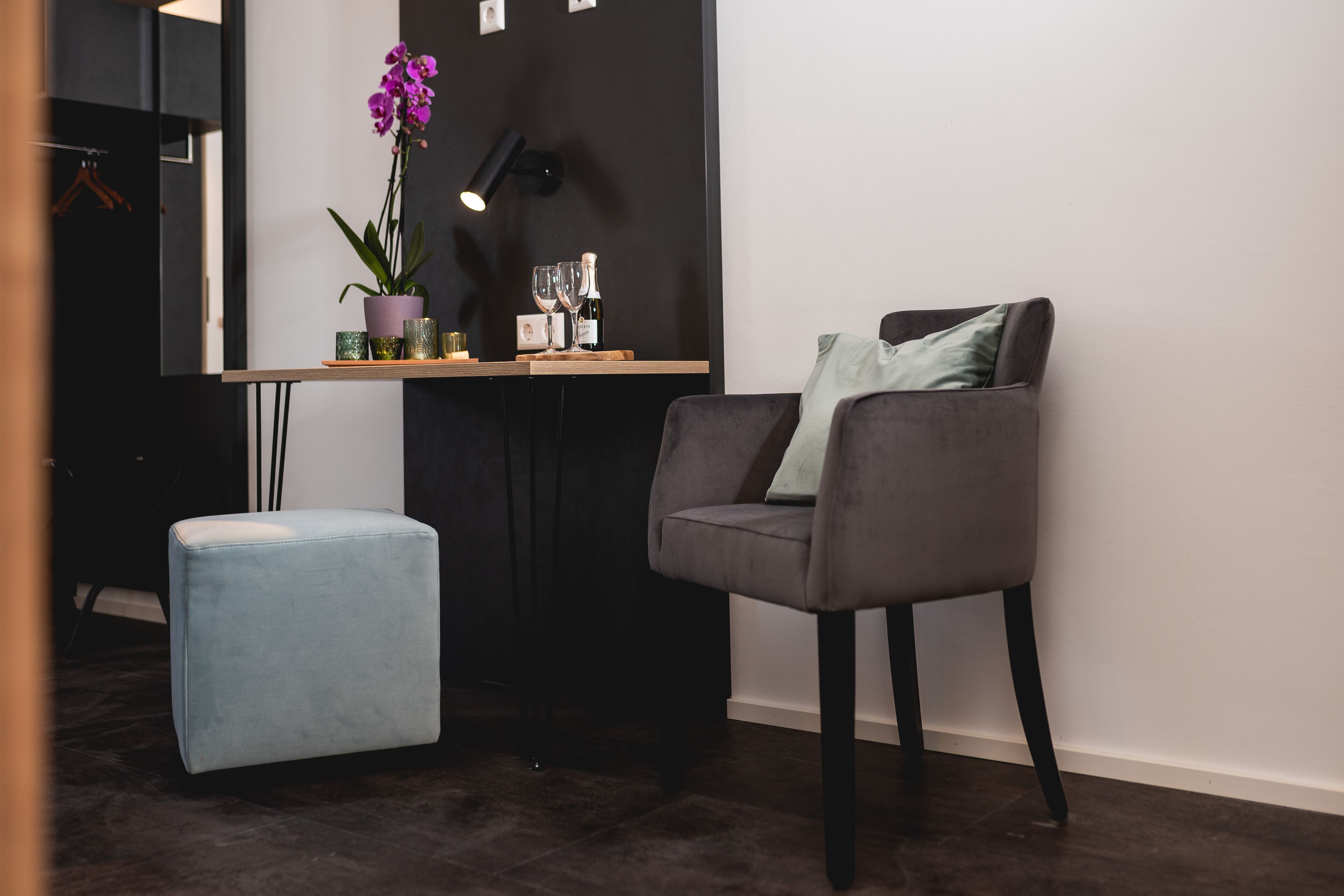 A modern room with a grey armchair, a small blue ottoman, a potted orchid, and a table with wine glasses and a bottle.