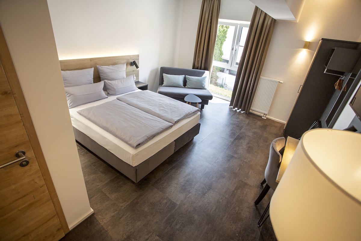 A modern hotel room with a large bed, a sofa, a desk, and a view of the outdoors.
