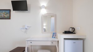 A bedroom with a bed, a desk, a chair, a small table, a mirror, a television, a refrigerator, and a wall socket. - Villa Stella (Santorini)