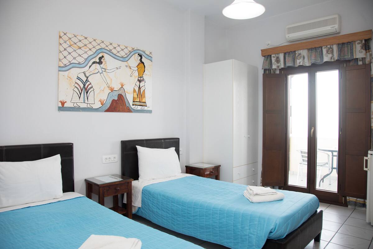 A hotel room with two beds, a large window with curtains, a painting on the wall, and a ceiling fan.