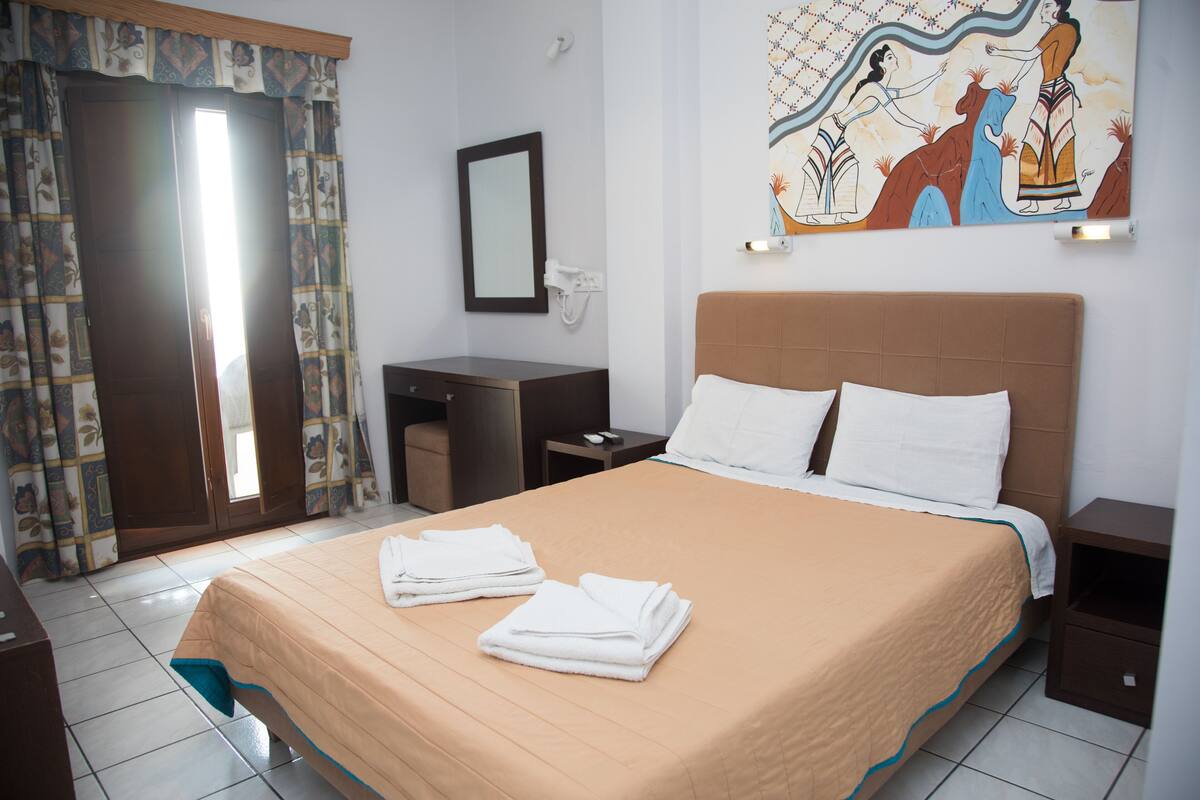 A hotel room with a bed, a desk, a chair, a nightstand, a mirror, and a painting on the wall.