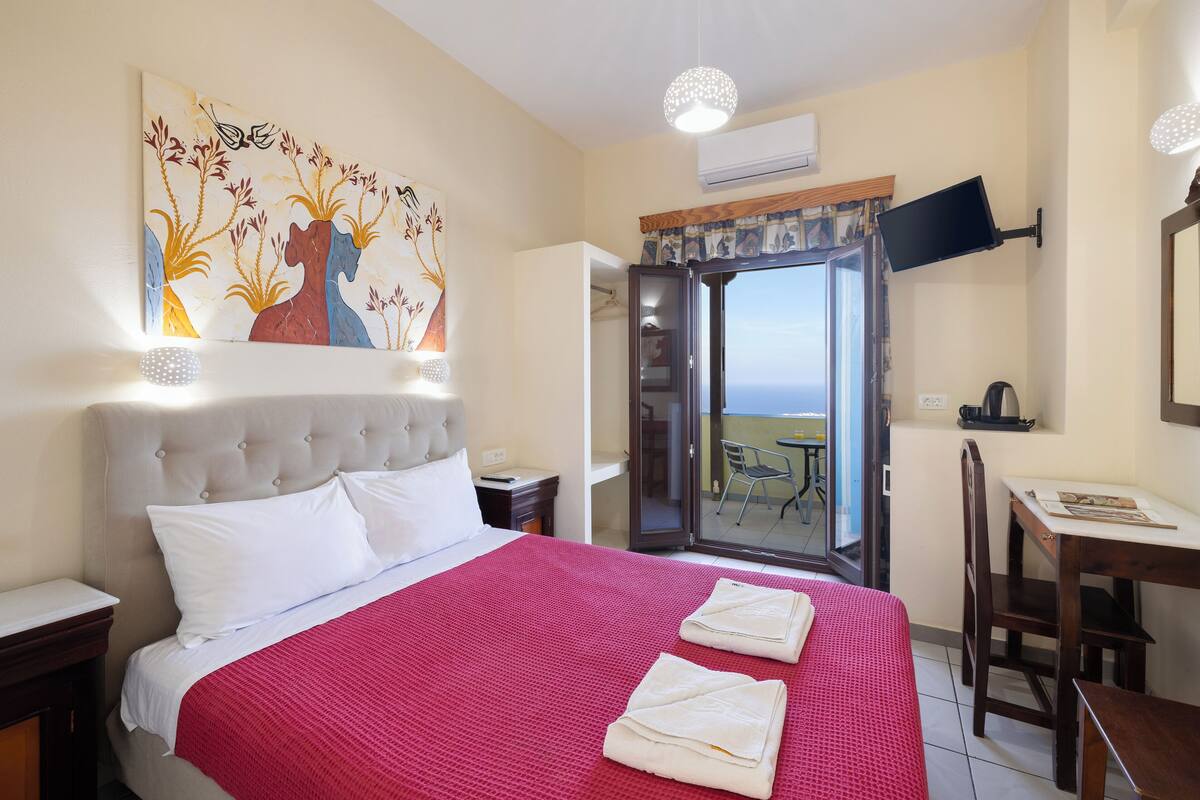 A hotel room with a large bed, a flat-screen TV mounted on the wall, a desk with a chair, and a balcony with a table and chairs.
