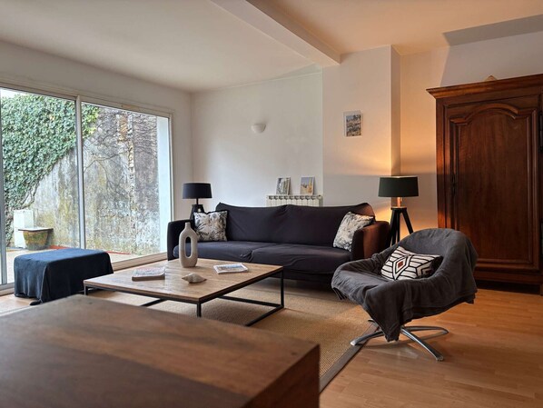 A modern living room with a sofa, coffee table, and armchair, large windows, and a wooden cabinet.