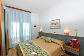 A hotel room with a bed, a nightstand, a lamp, a framed picture, and a bouquet of flowers. - Hotel Souvenir (Jesolo)
