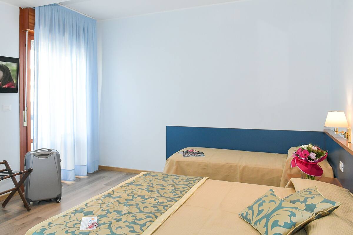A hotel room with a bed, a chair, a suitcase, and a table with a lamp and flowers.