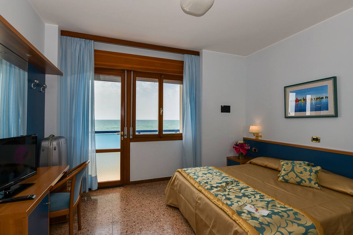 A hotel room with a bed, a desk with a television, a suitcase, and a view of the sea through a large window.