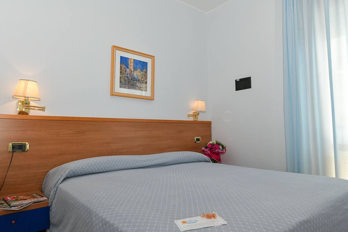 A hotel room with a neatly made bed, two bedside lamps, a framed picture on the wall, a small nightstand with a magazine, and a window with curtains.