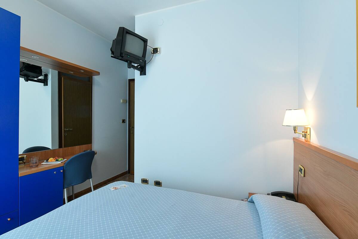 A hotel room with a bed, a desk with a chair, a television mounted on the wall, and a bedside lamp.