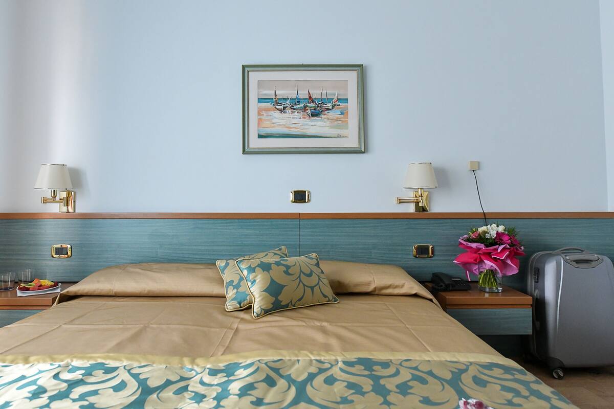 A hotel room with a bed, two bedside tables, a nightstand, a lamp, a painting, a vase with flowers, and a suitcase.