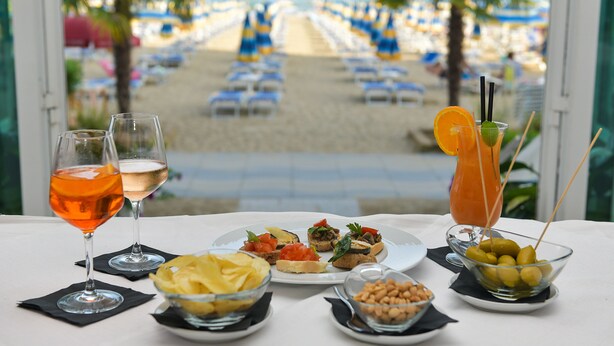 A table with drinks and appetizers, a view of a beach with rows of chairs, and a palm tree. - Hotel Souvenir (Jesolo)