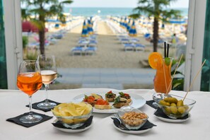 A table with drinks and appetizers, a view of a beach with rows of chairs, and a palm tree. - Hotel Souvenir (Jesolo)
