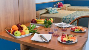 A hotel room with a wooden table set for breakfast, including fruits and plates of food. There are two beds with blue and beige bedding, and a nightstand with a lamp and flowers. - Hotel Souvenir (Jesolo)