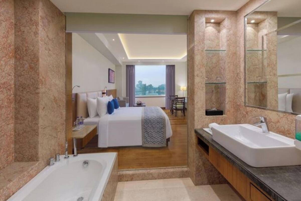 Habitación de hotel moderna con una cama grande, baño separado con bañera, zona de comedor y vistas al exterior.