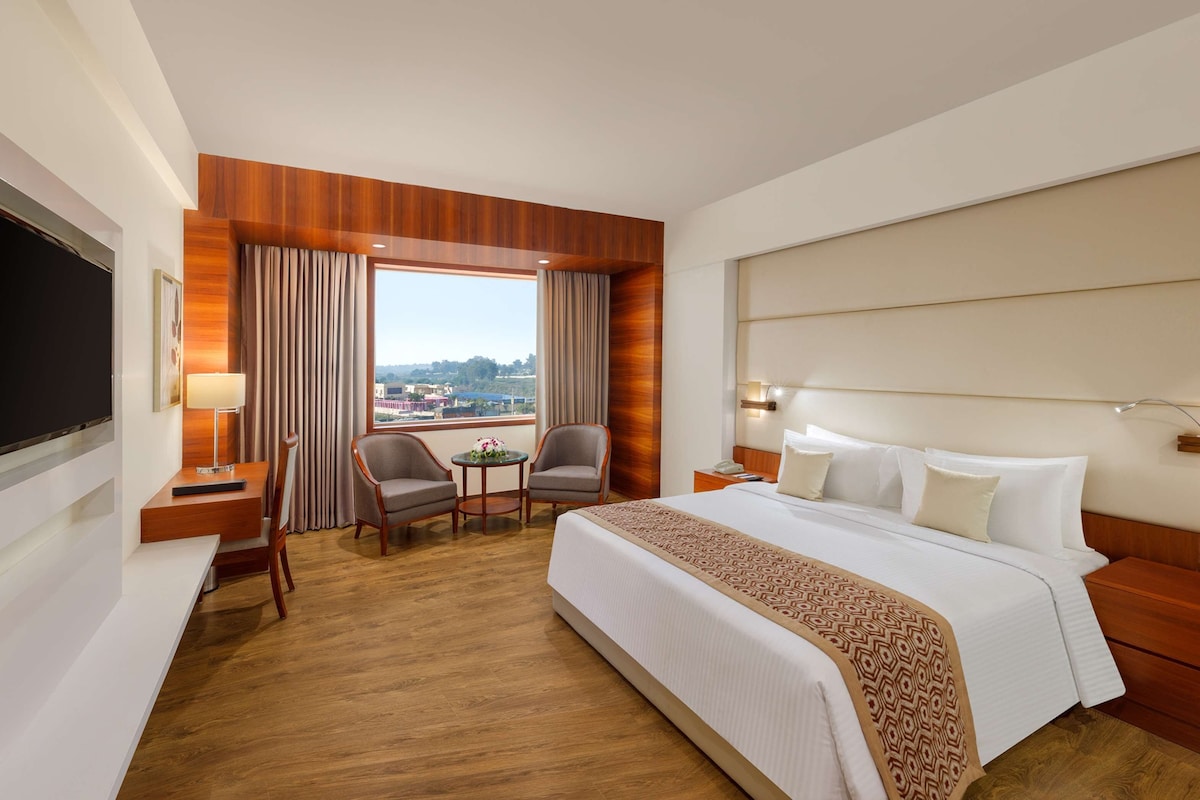 Habitación de hotel con una cama grande, un escritorio con televisor, dos sillones, una mesita con flores y vistas a la ciudad a través de amplios ventanales.