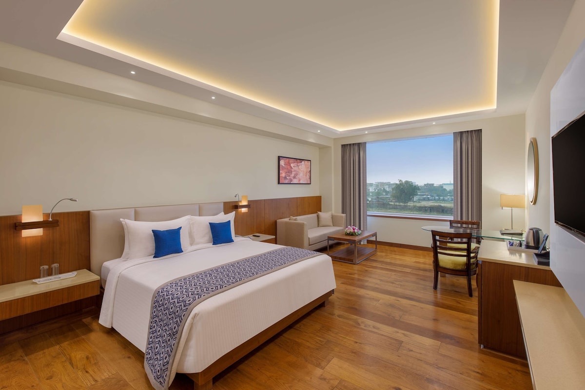 Habitación de hotel con una cama grande, un escritorio, una silla, un televisor y una ventana con vista a la ciudad.