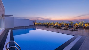 Piscina infinita en la azotea con sillones de descanso y palmeras, rodeada por un edificio moderno.