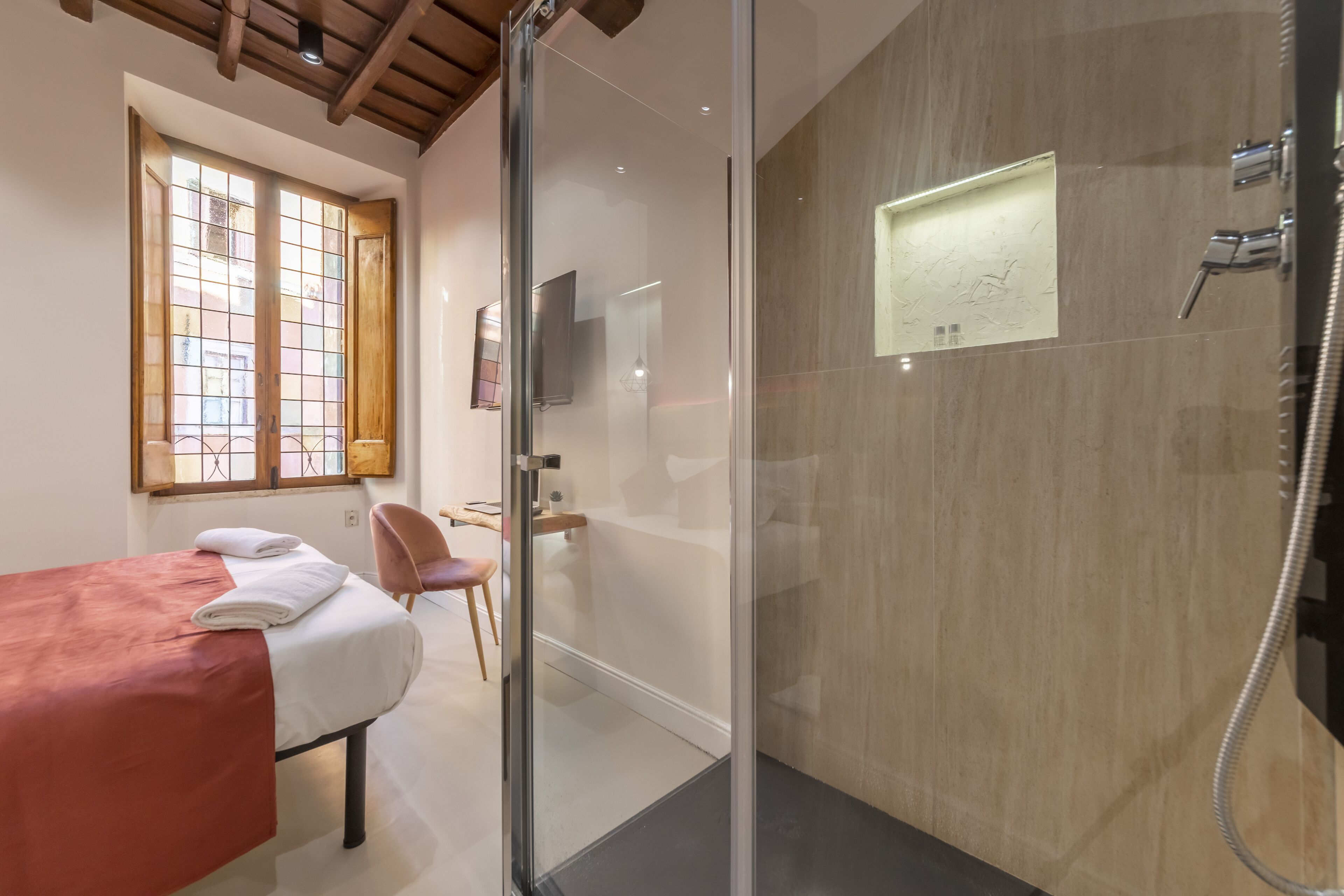 Une chambre d’hôtel moderne avec une douche en verre, un lit recouvert d’une couverture rouge, une chaise et un bureau.