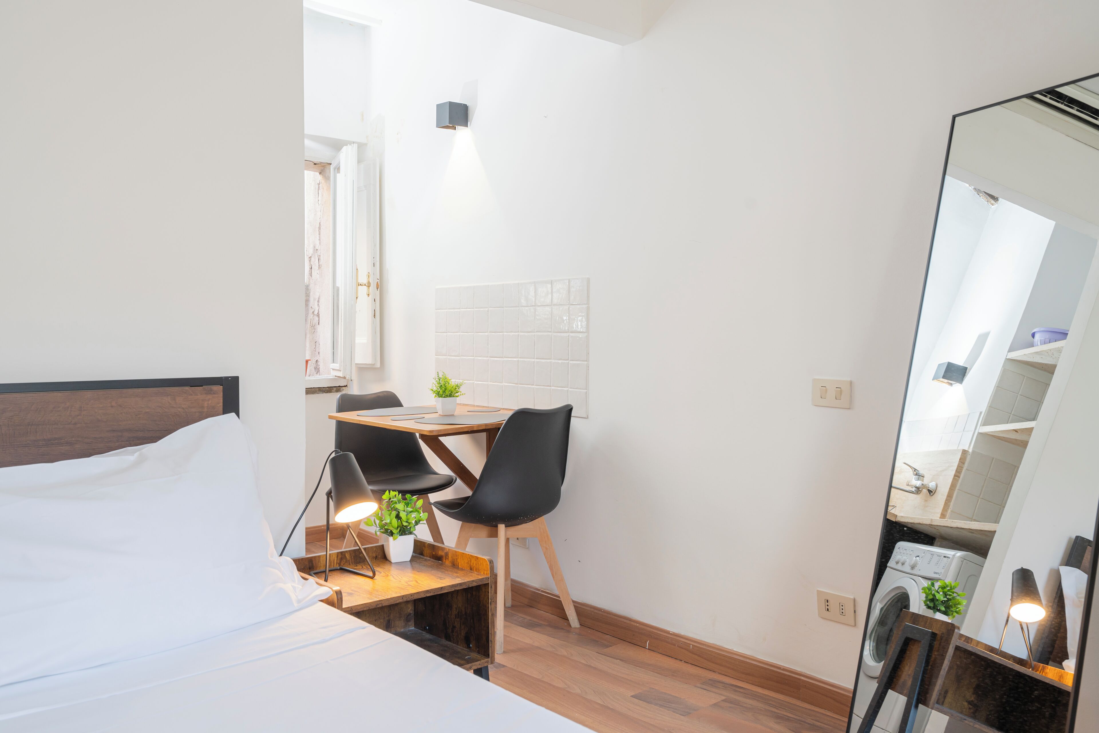 A compact hotel room with a bed, a small dining table with chairs, a mirror, and a washing machine.