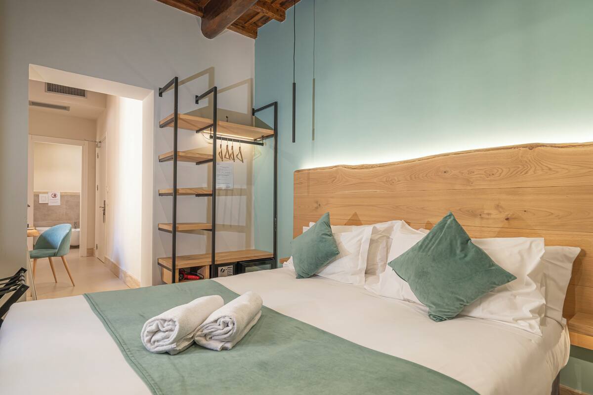 A neatly made bed with green bedding and pillows, a wooden headboard, and a wall-mounted shelf with hanging items.