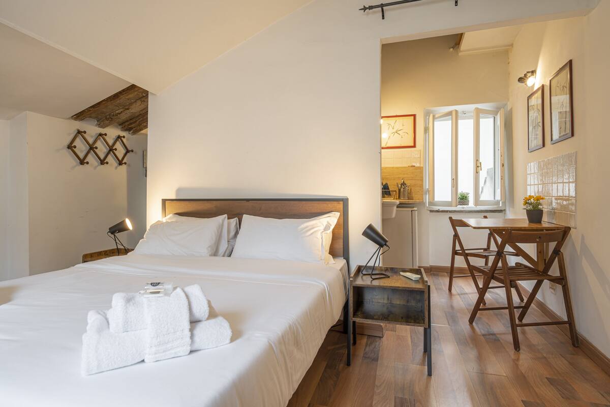 A hotel room with a large bed, bedside tables, a desk with chairs, and a view of the kitchen area.