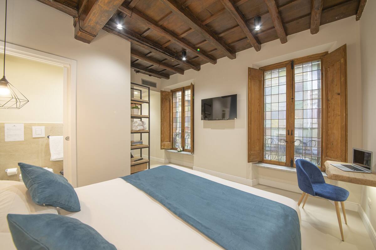 A bedroom with a large bed, a desk with a laptop, a chair, a TV, and a window with a view.