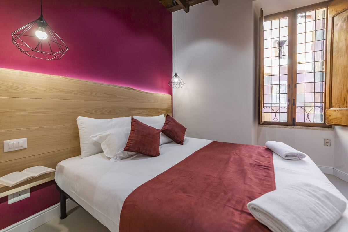 A neatly made bed with a wooden headboard, a red blanket, and two pillows. There is a window with bars and a wooden door to the right.