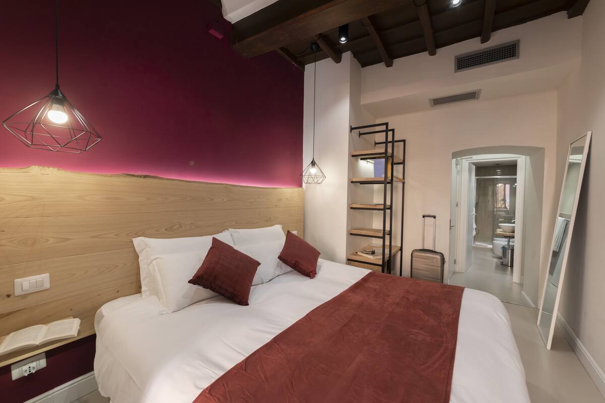 A modern hotel room with a wooden headboard, a large bed with white and brown bedding, a red accent throw, and a pendant light.