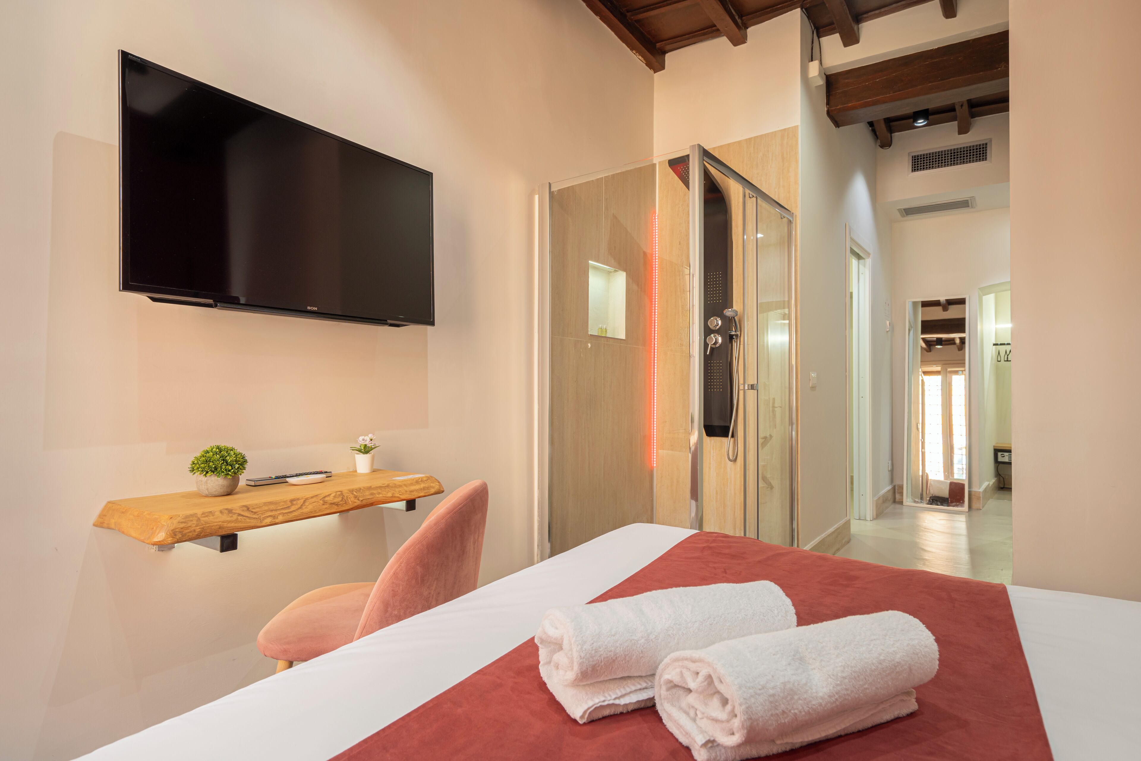 A hotel room with a bed, a TV mounted on the wall, a wooden shelf with a plant, and a shower area.