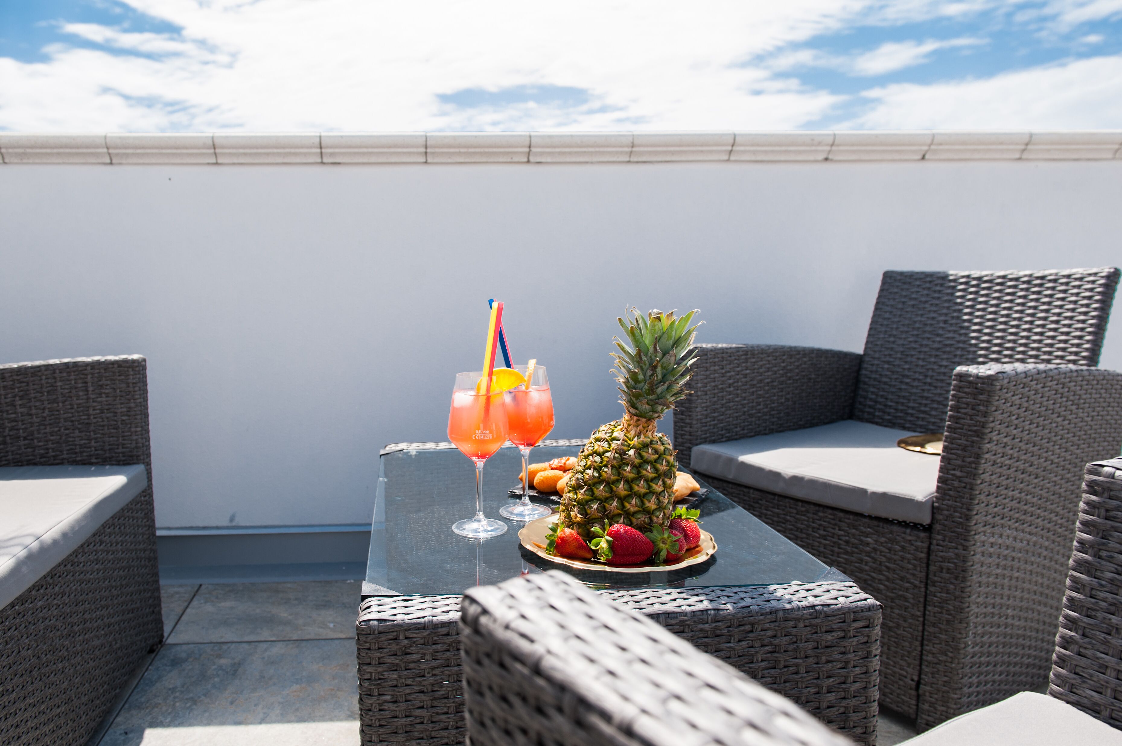Una terraza en la azotea con mobiliario de mimbre, una mesa con bebidas y frutas, y un cielo despejado.