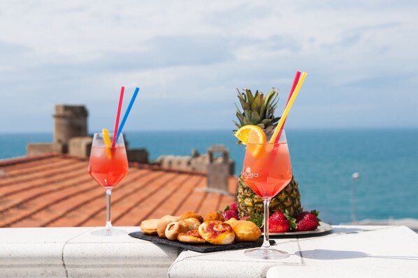 Two cocktails with colorful straws and a pineapple on a table with pastries, overlooking a coastal view.