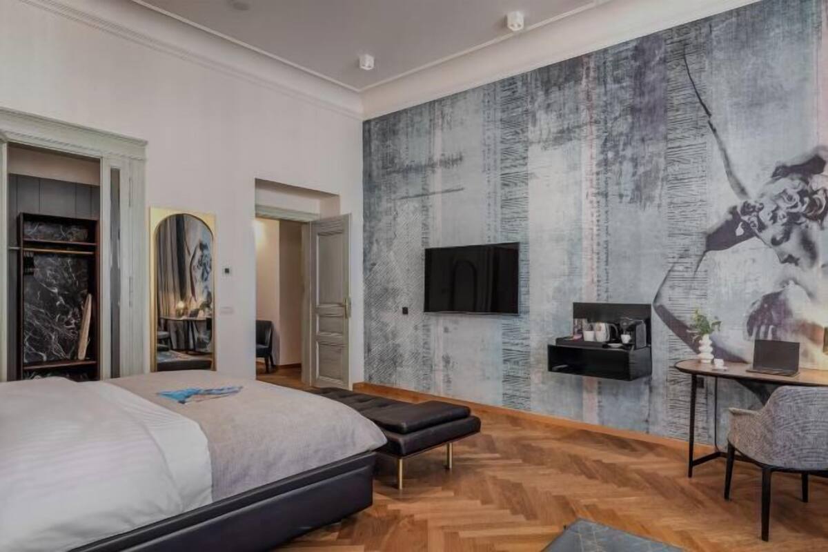 A modern bedroom with a large bed, a desk with a laptop, a TV mounted on the wall, and a large abstract painting.