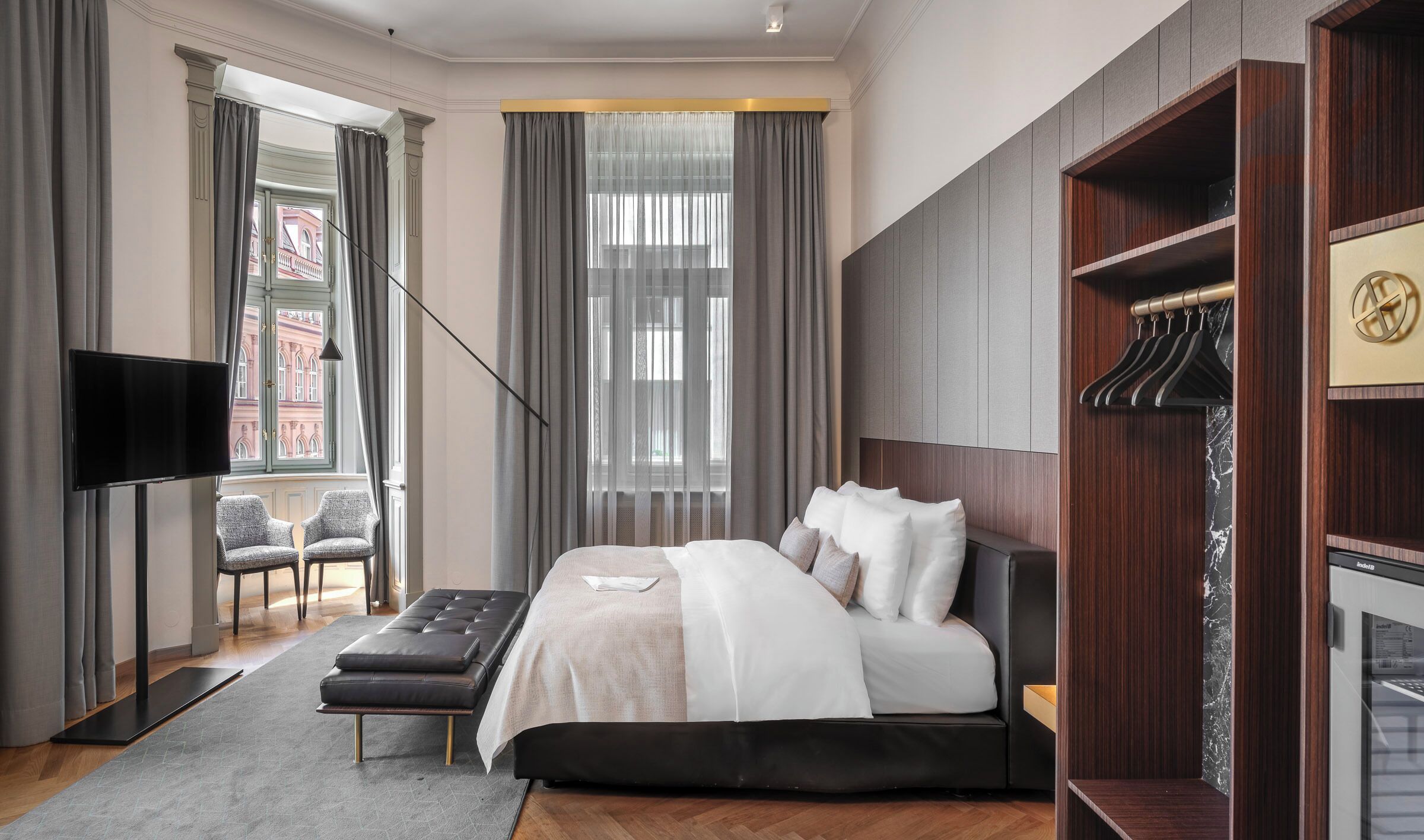 A modern hotel room with a large bed, a TV, a seating area with chairs, and a wardrobe with a mirror.